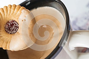 Sugared jam ring biscuit being dunked into frothy coffee in a mug