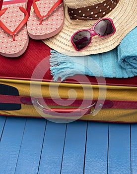 Suitcase with flip-flops, beach towel and sunglasses on wooden table.