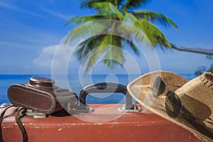 Suitcase, sun hat, photo camera and sunglasses with sea water, coconut palm tree and blue sky background on sunny summer day in