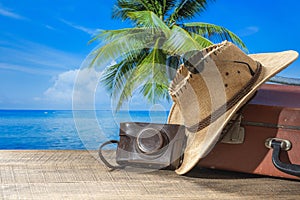 Suitcase, sun hat, photo camera and sunglasses on wooden deck with sea water, coconut palm tree and blue sky background on sunny