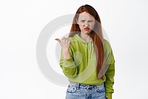 Sulking teen girl with red hair, grimacing and cringe reluctant, pointing finger left at something unfair or upsetting