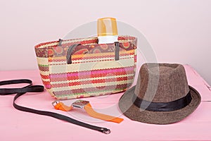 Summer accessories - straw beach bag, sun hat, belt, watch, suntan lotion, orange denim shorts on pink wooden table