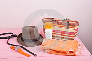 Summer accessories - straw beach bag, sun hat, belt, watch, suntan lotion, orange denim shorts on pink wooden table