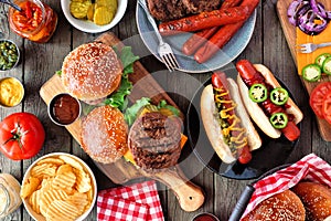 Summer BBQ food table scene with hot dog and hamburger buffet, top view over dark wood