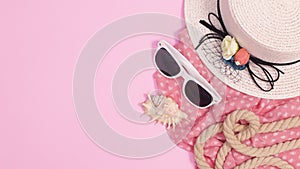 Summer beach hat, sunglasses and scarf appear with shells and rope on pastel pink background. Stop motion