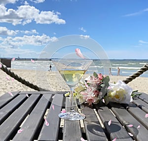 summer beach romantic flowers a bouquet , glass of wine on wooden table,in air  flying pink petal  view to sea at horizon blue sk