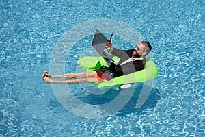 Summer business dreams. Business man in suit floating with cocktail and laptop in swimming pool. Summer business