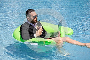 Summer business dreams. Business man in suit floating with cocktail and laptop in swimming pool. Summer business