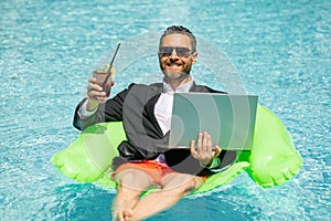 Summer business dreams. Millennial business man in suit floating with cocktail and laptop in swimming pool. Summer