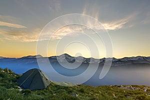 Summer camping in mountains at dawn. Tourist tent on round grassy hill on distant misty blue mountains range under pink sky before