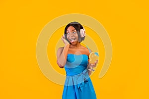 Summer clubbing. Black lady with tropical cocktail wearing headphones, listening to dance music on orange background