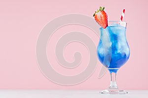 Summer cold blue lagoon alcohol cocktail with curacao liquor, strawberry slice, ice cubes, straw in misted glass on pink.