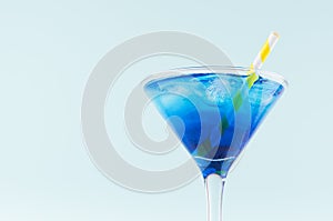 Summer cold blue lagoon drink with ice cube, yellow striped straw in elegant martini glass on pastel mint background, closeup.