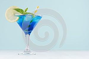 Summer cold blue lagoon drink with ice cubes, lemon slice, yellow straw, green mint in elegant wine glass on pastel mint.