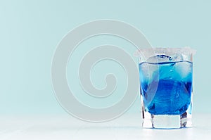 Summer cold blue lagoon drink with ice cubes, salt rim in elegant shot glass on pastel mint background.