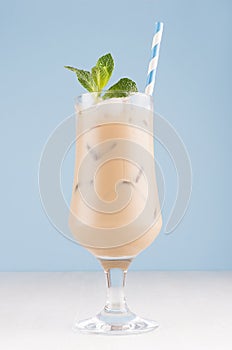 Summer cold coffee cocktail with cream, ice cubes, striped straw, green mint in transparent glass in soft pastel blue interior.