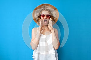 Summer concept. crazy girl in a sun hat and glasses shouts on a blue isolated background, a woman speaks loudly in summer clothes