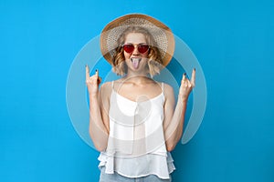 Summer concept. crazy girl in a sun hat and glasses shows rocknrol sign on a blue isolated background, a woman in summer clothes