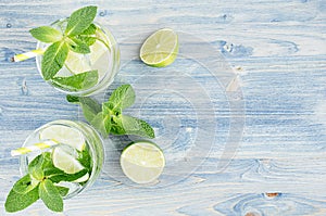 Summer cool lemonade in two wet glasses with mint, lime, ice, straw on soft shabby blue wood board, copy space, top view.