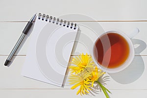 Summer cute flat lay: a cup of tea, a notepad with a blank page, a pen and three yellow daisies.