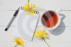 Summer cute flat lay: a cup of tea, a notepad with a blank page, a pen and three yellow daisies.
