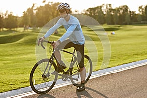 Summer cycling training. Young athletic man in sportswear riding bicycle along a road in city park at sunset, full