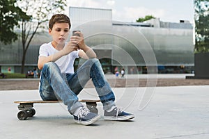 Summer day. Boy in white T-shirt and jeans sitting outdoor on longboard and uses smartphone. Boy plays computer games