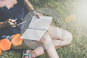 Summer day.Close-up of smartphone and notepad in hand of young woman sitting in park on lawn and reading book.