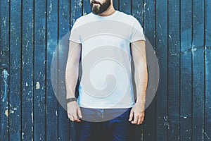 Front view. Young bearded hipster man dressed in white t-shirt and sunglasses is stands against dark wood wall.