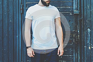 Front view. Young bearded hipster man dressed in white t-shirt and sunglasses is stands against dark wood wall.