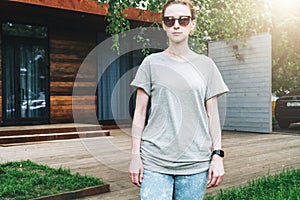 Front view. Young millennial woman dressed in gray t-shirt and sunglasses is stands on city street. Mock up.