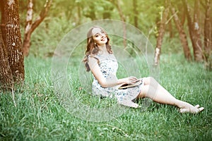 Summer day portrait of a beautiful cute young woman or girl sitting in a dress on the grass in a park with a book in her arms, sm