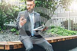 Summer day. Young bearded businessman in suit and tie sitting in park on bench, holding laptop and using smartphone