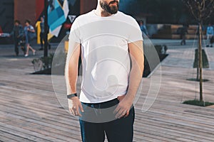 Summer day. Young bearded hipster man dressed in white t-shirt and sunglasses is stands on city street. Mock up