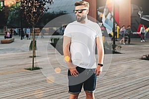 Summer day. Young bearded hipster man dressed in white t-shirt and sunglasses is stands on city street. Mock up