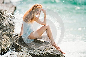 Summer dream. Sexy woman sitting on rocky beach against sea blue waves. Sensual young woman wearing sexy summer dress on