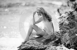 Summer dream. Sexy woman sitting on rocky beach against sea blue waves. Sensual young woman wearing sexy summer dress on