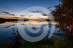 Summer evening twilight view on picturesque plain lake with forest and blossoming meadows on shores and sunset clouds reflections