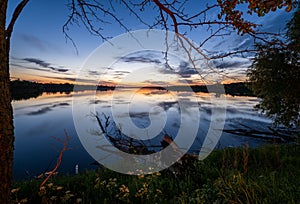 Summer evening twilight view on picturesque plain lake with forest and blossoming meadows on shores and sunset clouds reflections