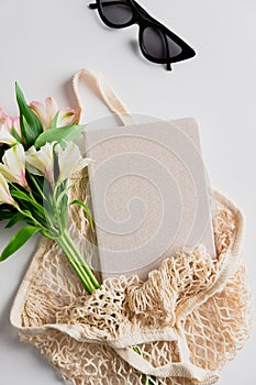 Summer fashion beige string bag with femenine accessories, bouquet of flowers, notebook Flat lay, top view