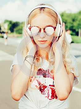 Summer fashion stylish portrait of young pretty blonde girl posing in sunglasses, T-shirt, and listening to music with headph