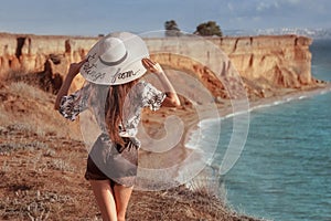 Summer fashionable girl portrait. Back view of trendy casual brunette woman in white hat resting on cliff mountains enjoying