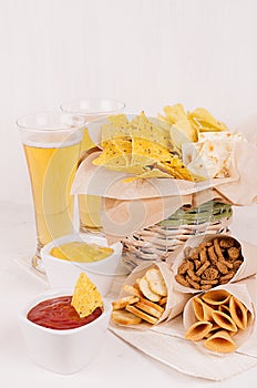 Summer fast food - different crunchy snacks and red, yellow sauce in white bowls, cold beer on soft white wood board, vertical.