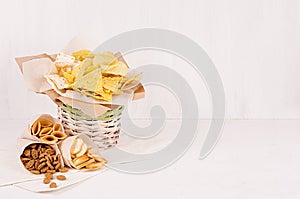 Summer fast food - different crunchy snacks in wicker rustic basket and craft paper cornet on soft white wood board.
