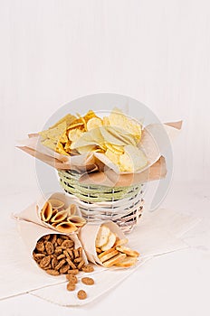 Summer fast food - different crunchy snacks in wicker rustic basket and craft paper cornet on soft white wood board, closeup.