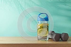 Summer fitness background with infused water bottle and dumbbells on wooden table over blue background with palm tree shadows