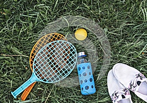 Summer flat lay on green grass with rackets and ball, glass bottle of water and summer white shoes