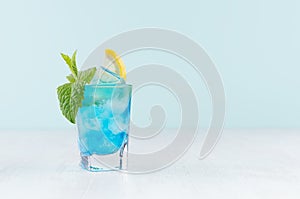 Summer fresh blue curacao cocktail with lemon slice, ice cubes, mint in wet shot glass on white wood table, pastel green.