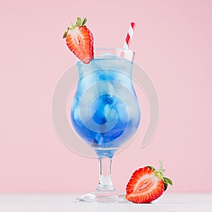 Summer fresh fruit blue curacao cocktail with strawberry slice, ice cubes, straw in wet glass on white wood table, pink background