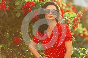 Summer fresh portrait of a beautiful young woman with fashionable sunglasses in a stylish red dress stands near a bush with